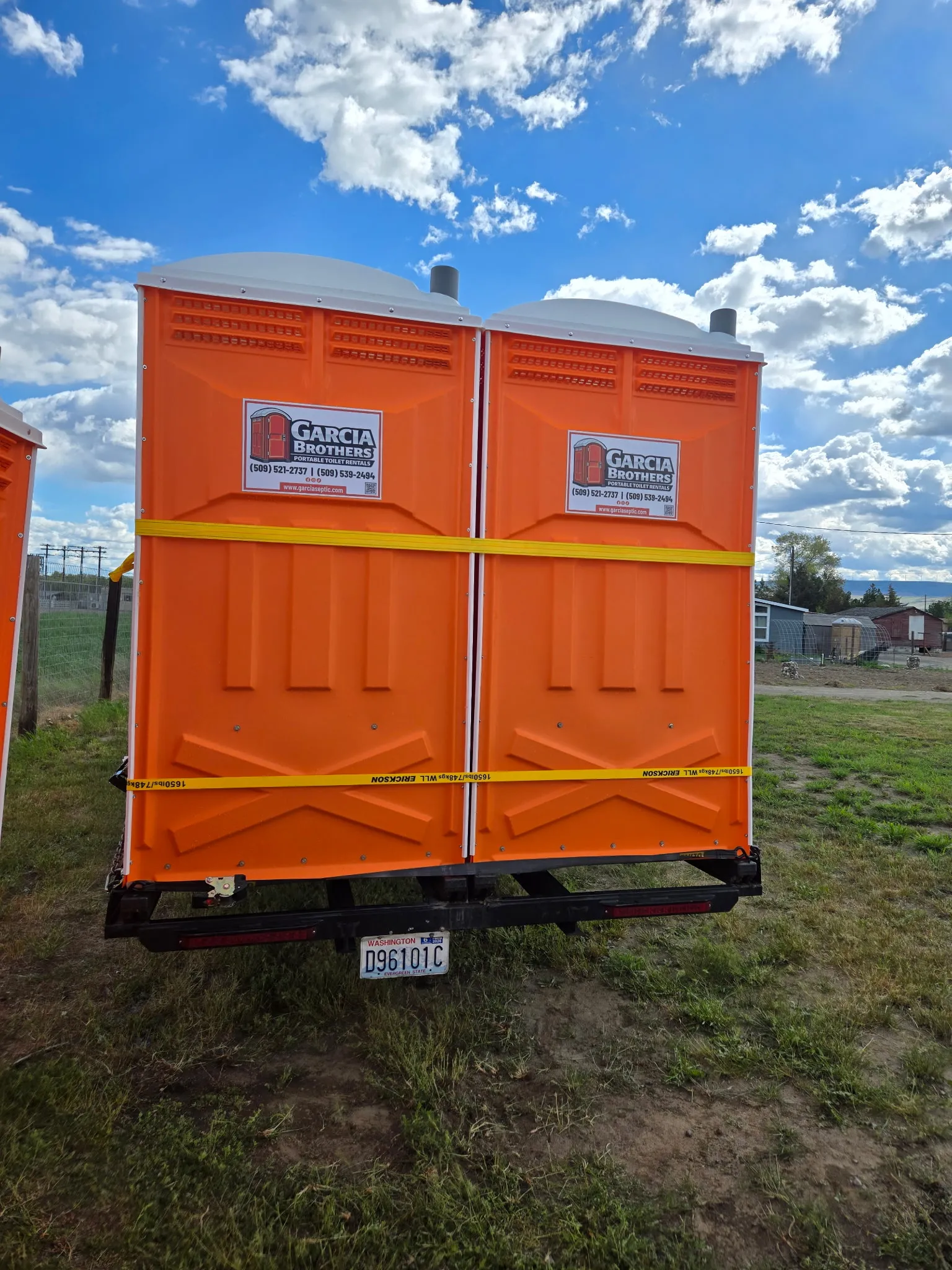 A pair of clean orange Garcia Brothers Portable Toilet Rentals units standing side by side in a grassy field under a blue sky, each marked with the company logo decal and contact numbers