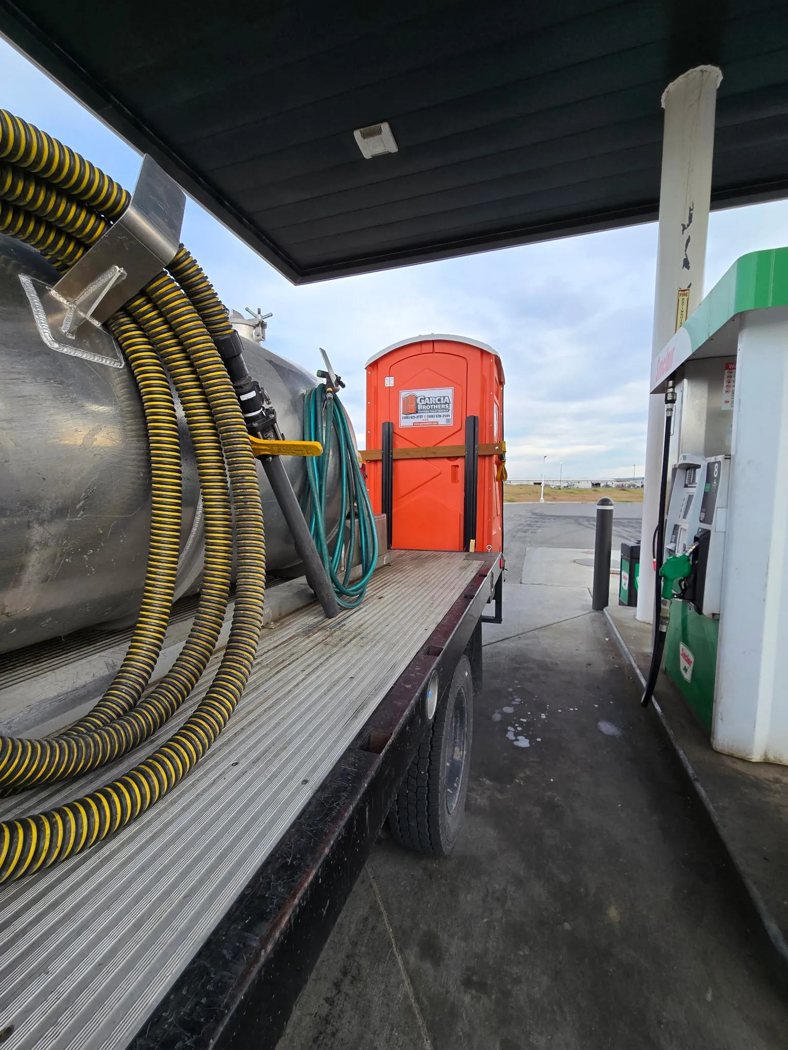 A Garcia Brothers flatbed service truck with its stainless tank and coiled hoses refueling at a station, an orange portable toilet loaded on the bed behind it
