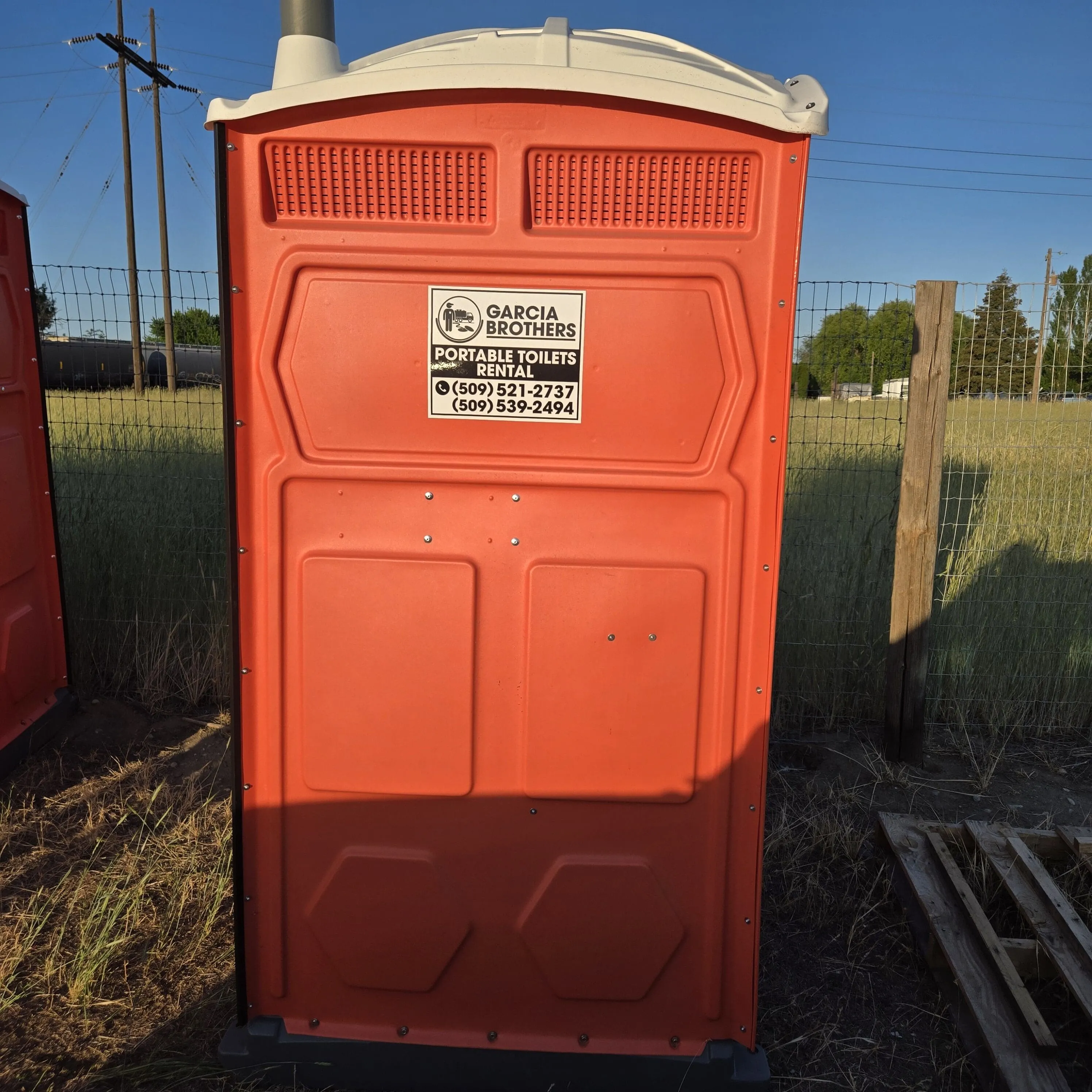 A clean orange Garcia Brothers portable toilet in a field, with the Portable Toilets Rental decal and contact numbers on the door