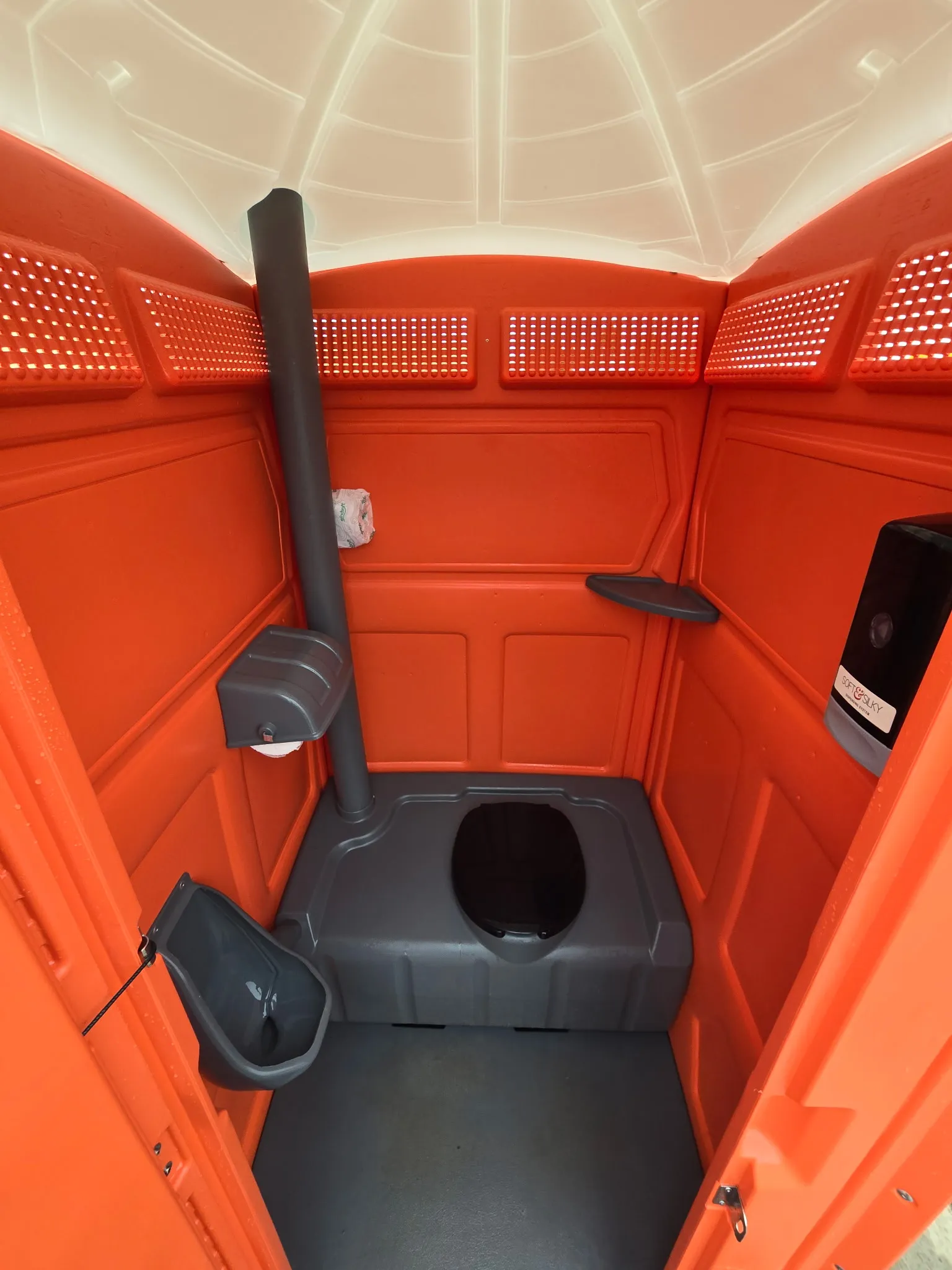 The clean interior of a standard Garcia Brothers portable toilet, showing the toilet, urinal, built-in hand sanitizer dispenser, and stocked toilet paper