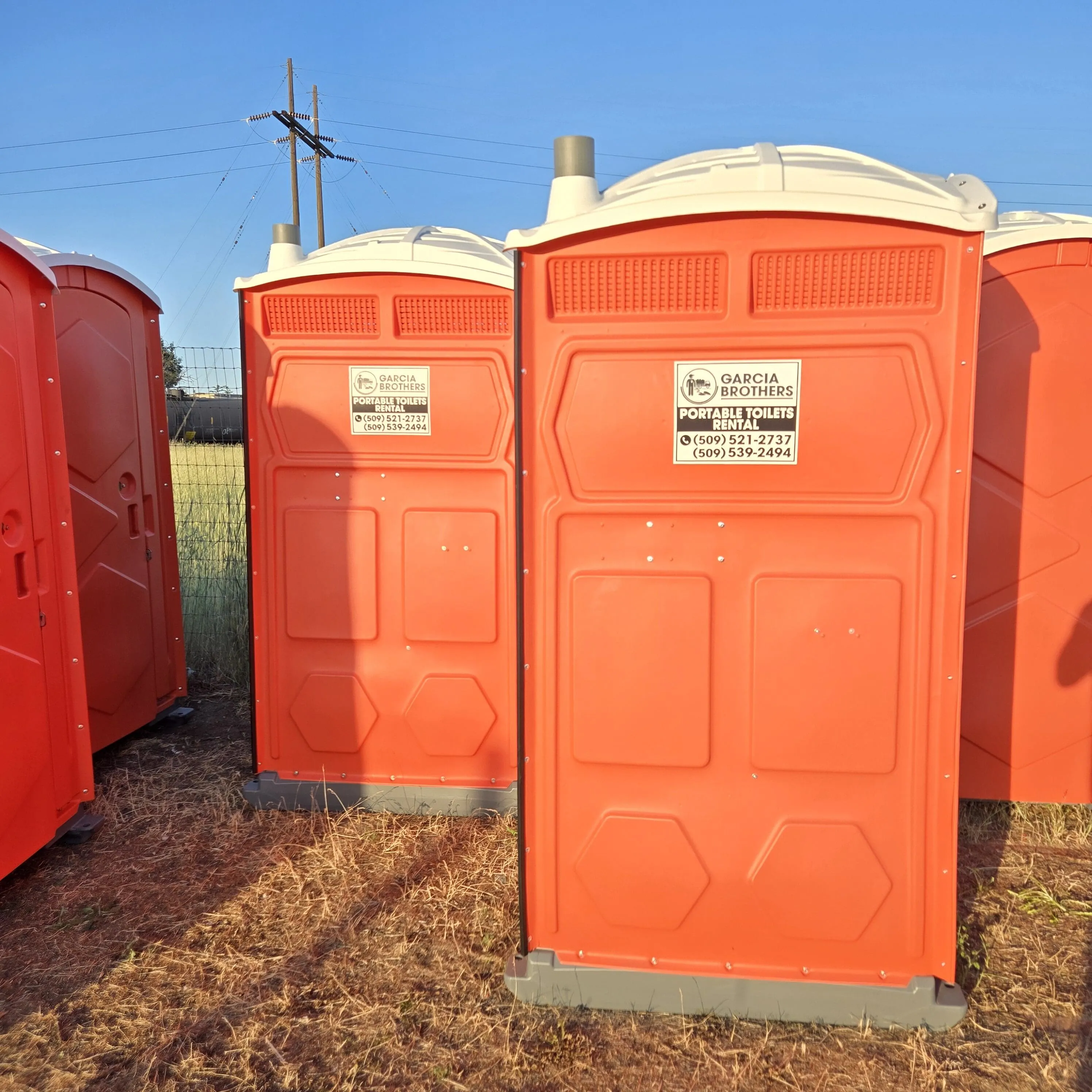 A row of orange Garcia Brothers portable toilets lined up in a field at golden hour, ready for delivery